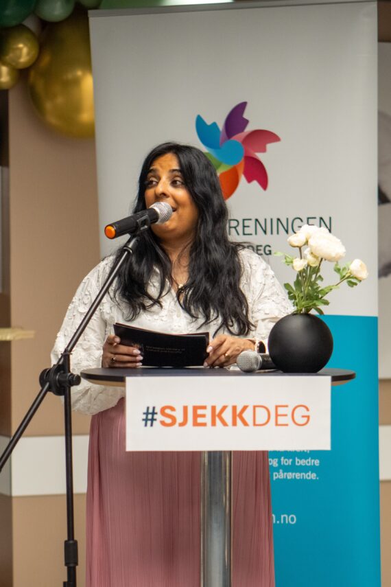 En kvinne, Lubna Jaffery, står på et podium merket #SJEKKDEG og snakker i en mikrofon. I bakgrunnen sees et banner og en vase med hvite blomster.