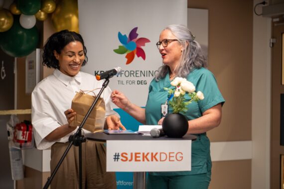 To kvinner står ved et #SJEKKDEG-podium under et arrangement. Den ene overrekker en brun papirpose til den andre, begge smiler. I bakgrunnen skimtes et banner og ballonger som omtaler livmorhalskreft.