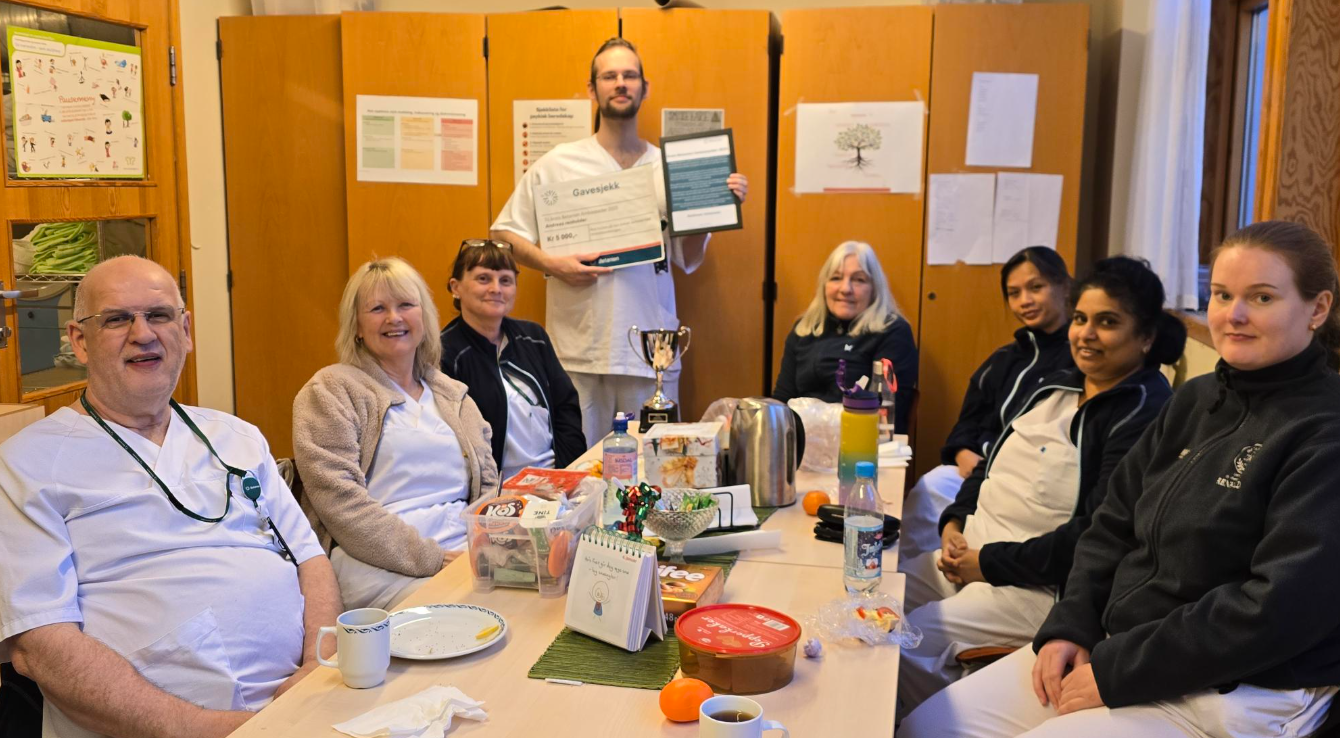 En gruppe på åtte helsearbeidere fra Betanien sitter rundt et bord med mat og drikke; en person står med et sertifikat og en plakett i hånden og feirer anerkjennelsen fra ambassadøren 2025.