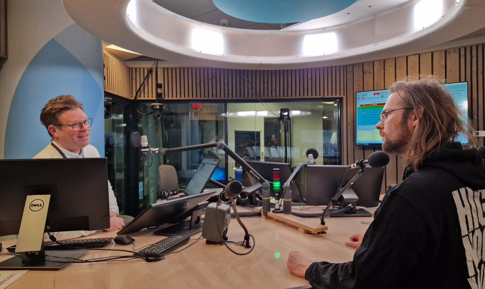 To personer sitter overfor hverandre ved en pult i et radiostudio med mikrofoner, datamaskiner og skjermer synlige i bakgrunnen mens de diskuterer