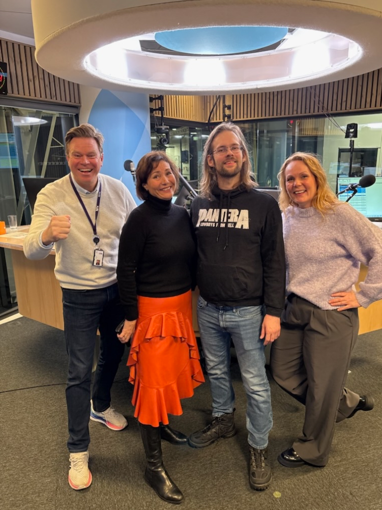 Fire personer står og smiler i et radiostudio, med mikrofoner og lydutstyr synlig i bakgrunnen