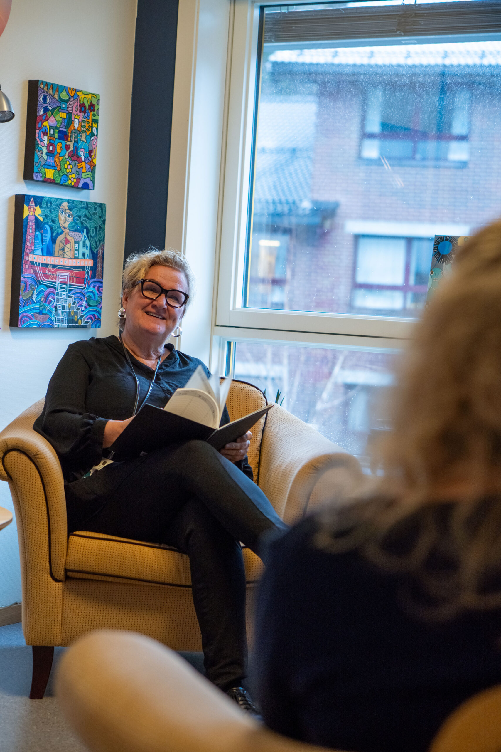 En kvinne sitter i en lenestol ved et vindu på Betanien sykehus, holder i en notatbok og smiler, mens hun nyter sine 25 år med jobb med mennesker. På veggen henger fargerike kunstverk, og i forgrunnen dukker en annen person opp.