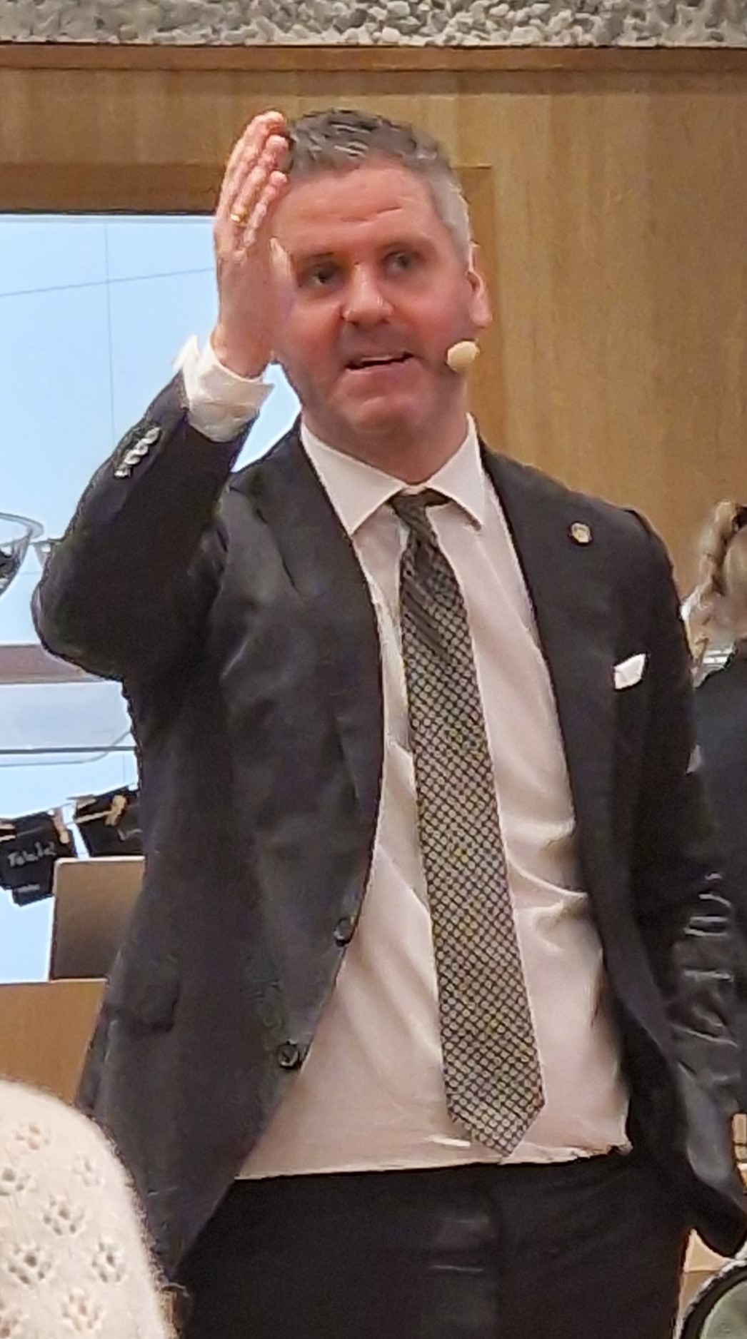 En mann i dress og slips snakker med en hodesettmikrofon og gestikulerer med løftet hånd i et moderne innemiljø