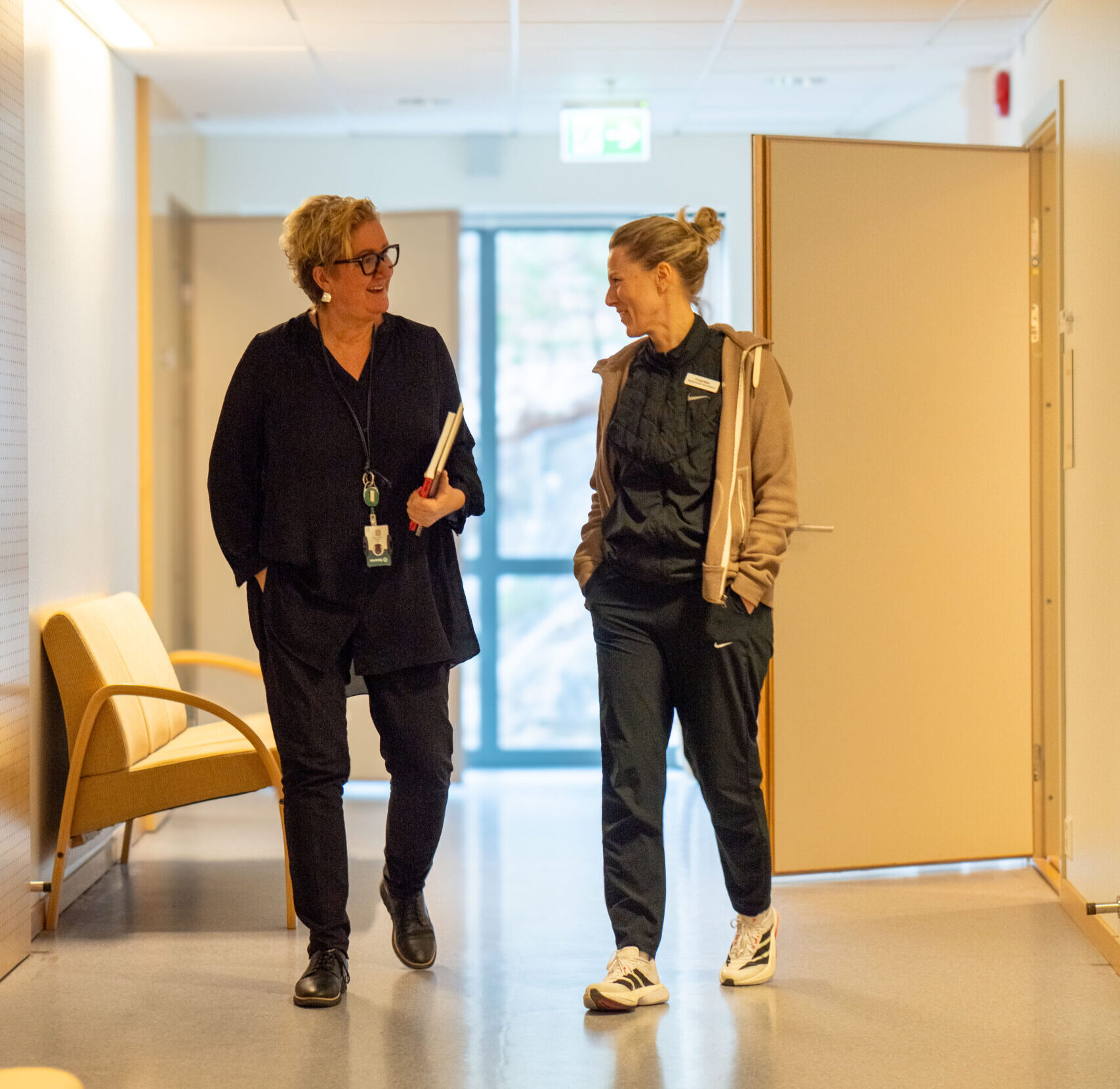 To kvinner går og snakker i en gang med lyse vegger på Betanien sykehus. Den ene holder en mappe.