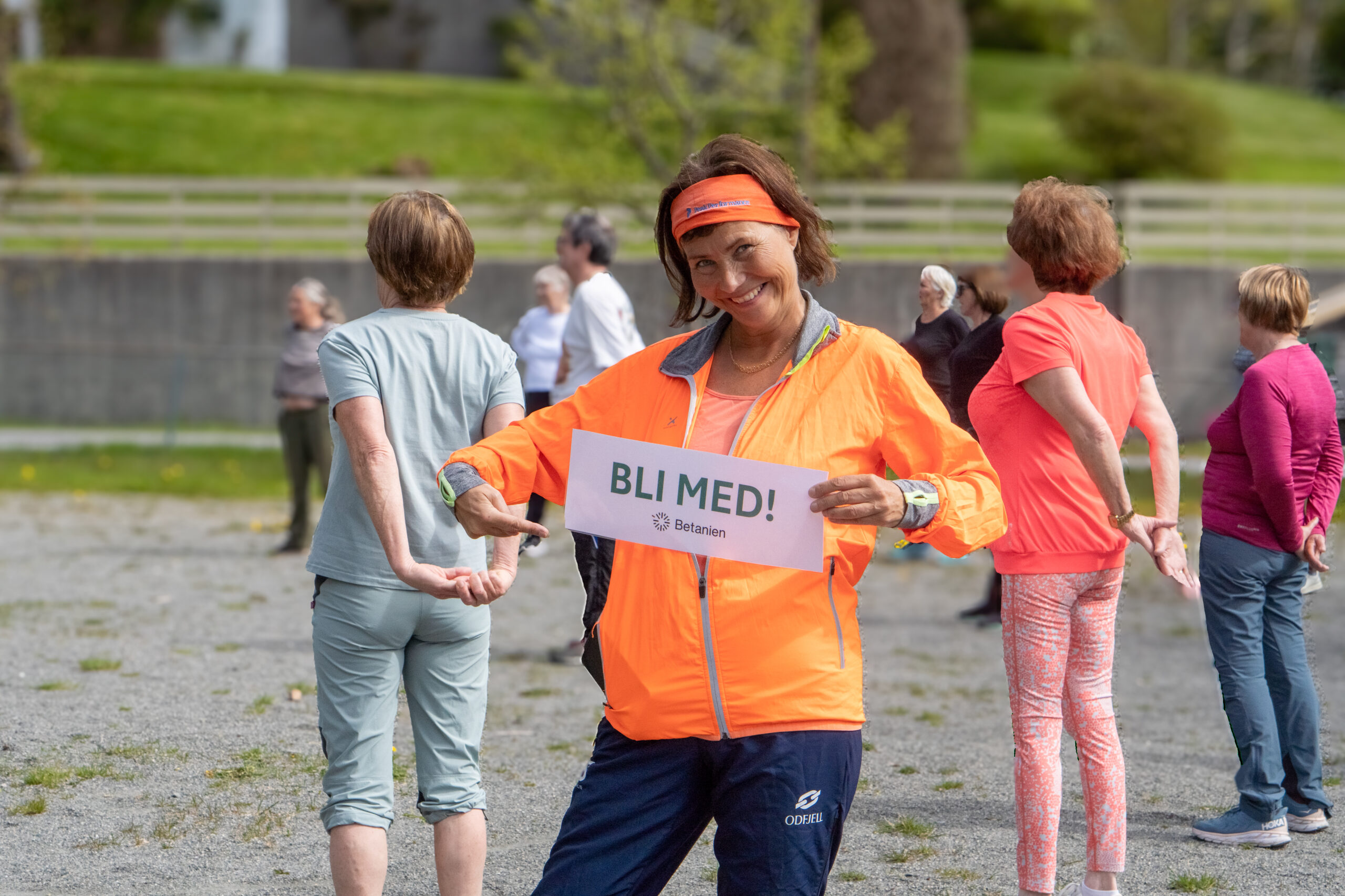 En kvinne i sportsklær smiler og peker på et skilt der det står BLI MED! mens andre trener utendørs i bakgrunnen.