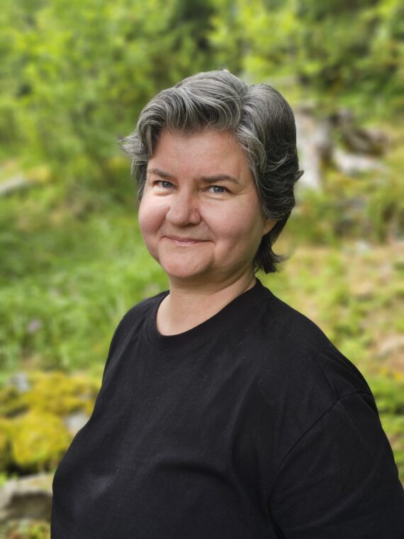 En person med kort, grått hår, kledd i en svart skjorte, står utendørs foran en grønn, uskarp naturbakgrunn.