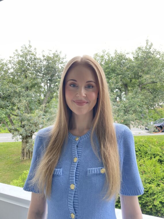 En kvinne med langt, rett hår, kledd i en blå genser med korte ermer og gullknapper, står ute foran grønne trær og busker.