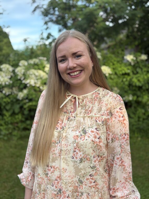 En kvinne med langt blondt hår, kledd i en lys bluse med blomstermønster, står og smiler ute i en hage med grønne planter og hvite blomster i bakgrunnen.