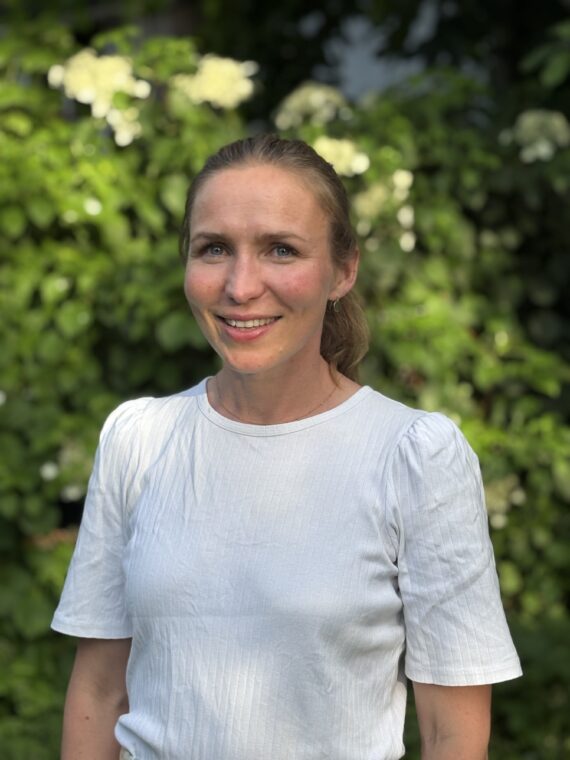 En kvinne med lysebrunt hår som er satt opp, står utendørs iført en hvit skjorte med korte ermer, med grønt løvverk og hvite blomster i bakgrunnen.