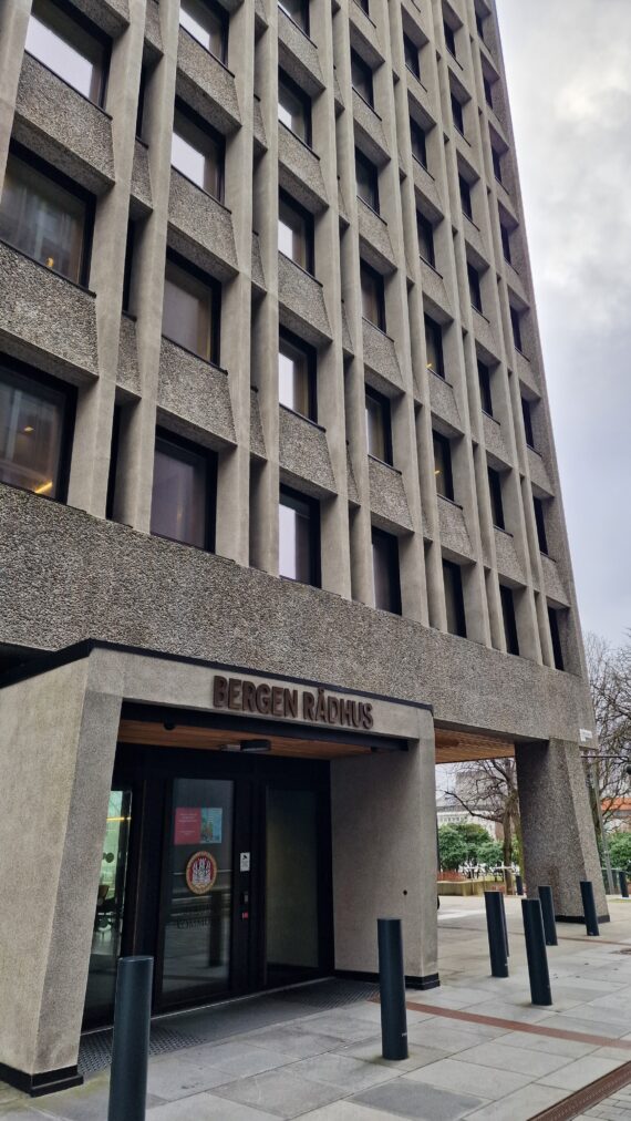 En høy betongbygning med mange vinduer, merket Bergen Rådhus over hovedinngangen, huser sentrale kontorer for Bergen kommune.