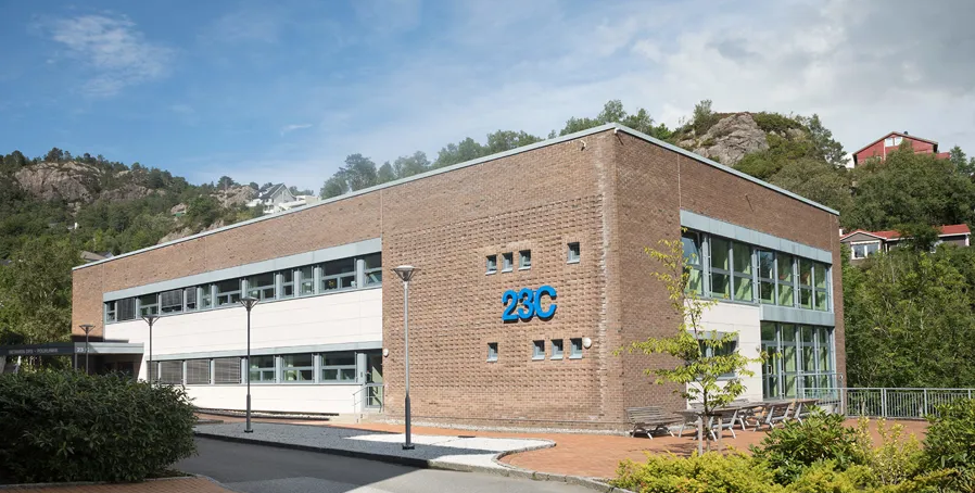 En toetasjes murbygning med store vinduer og 23C i blå bokstaver på utsiden, omgitt av grøntområder og små åser i bakgrunnen