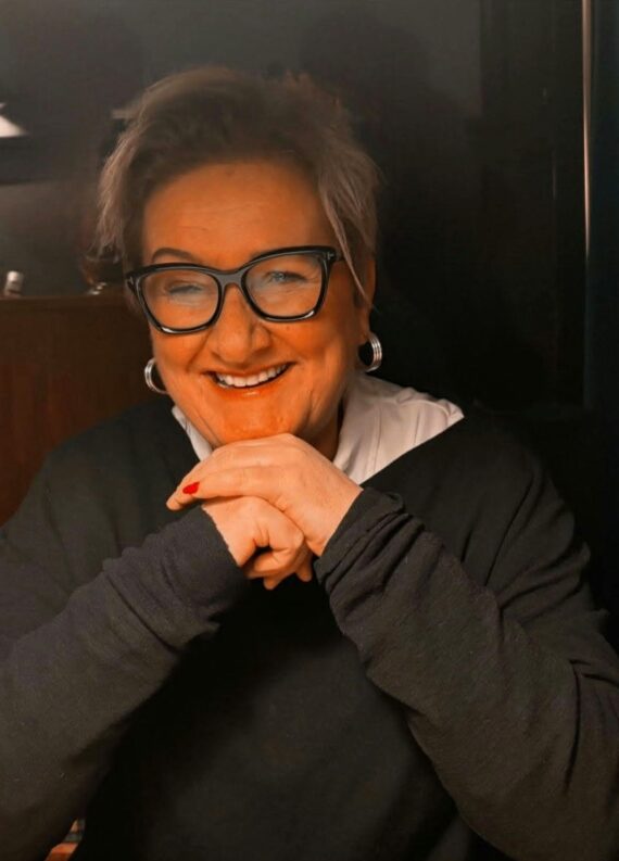 En kvinne med kort, grått hår, briller og øreringer sitter ved et bord og smiler med hendene foldet foran seg. .