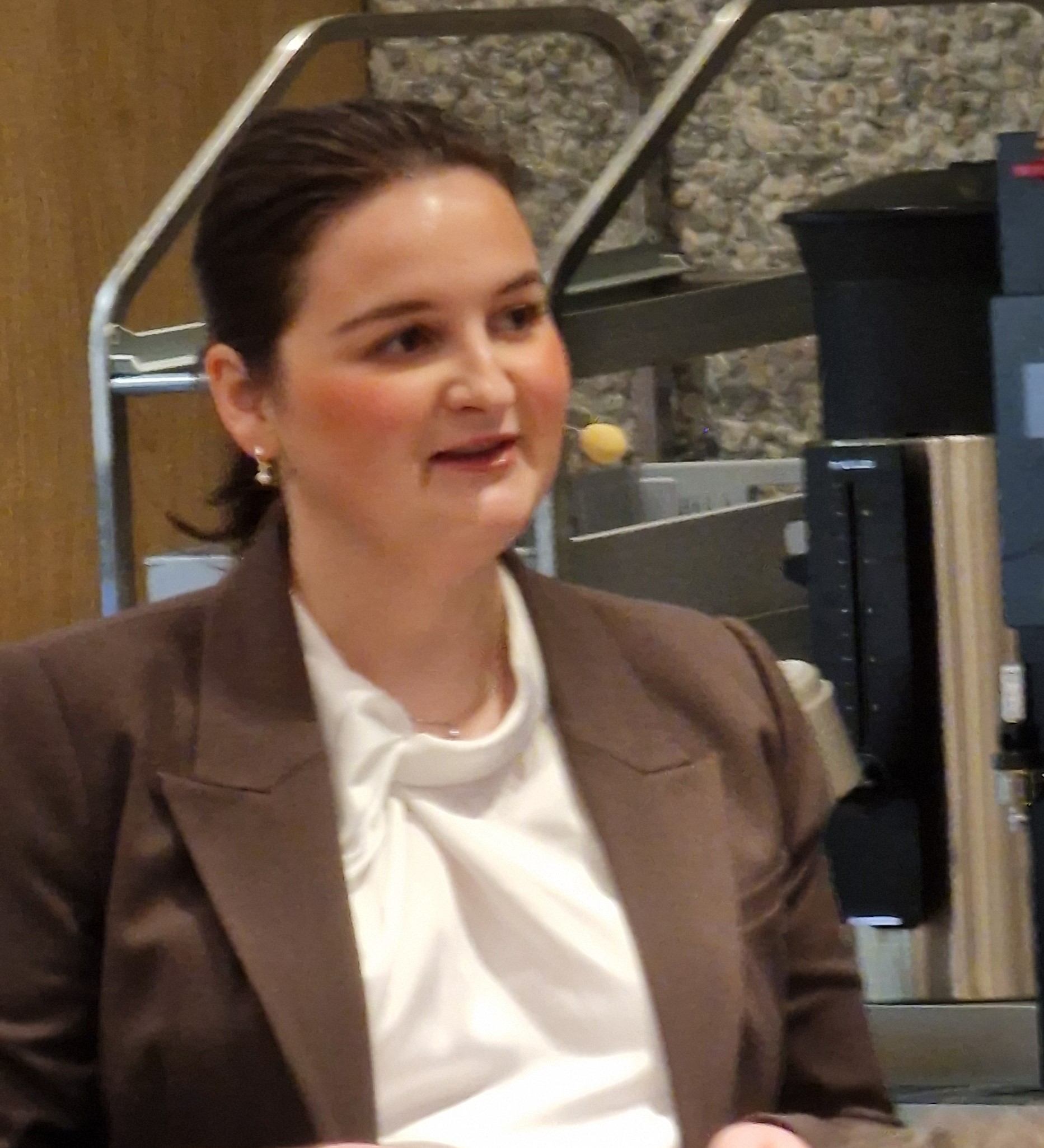 En kvinne med brunt hår, iført brun blazer og hvit bluse, snakker innendørs med et mikrofonhodesett