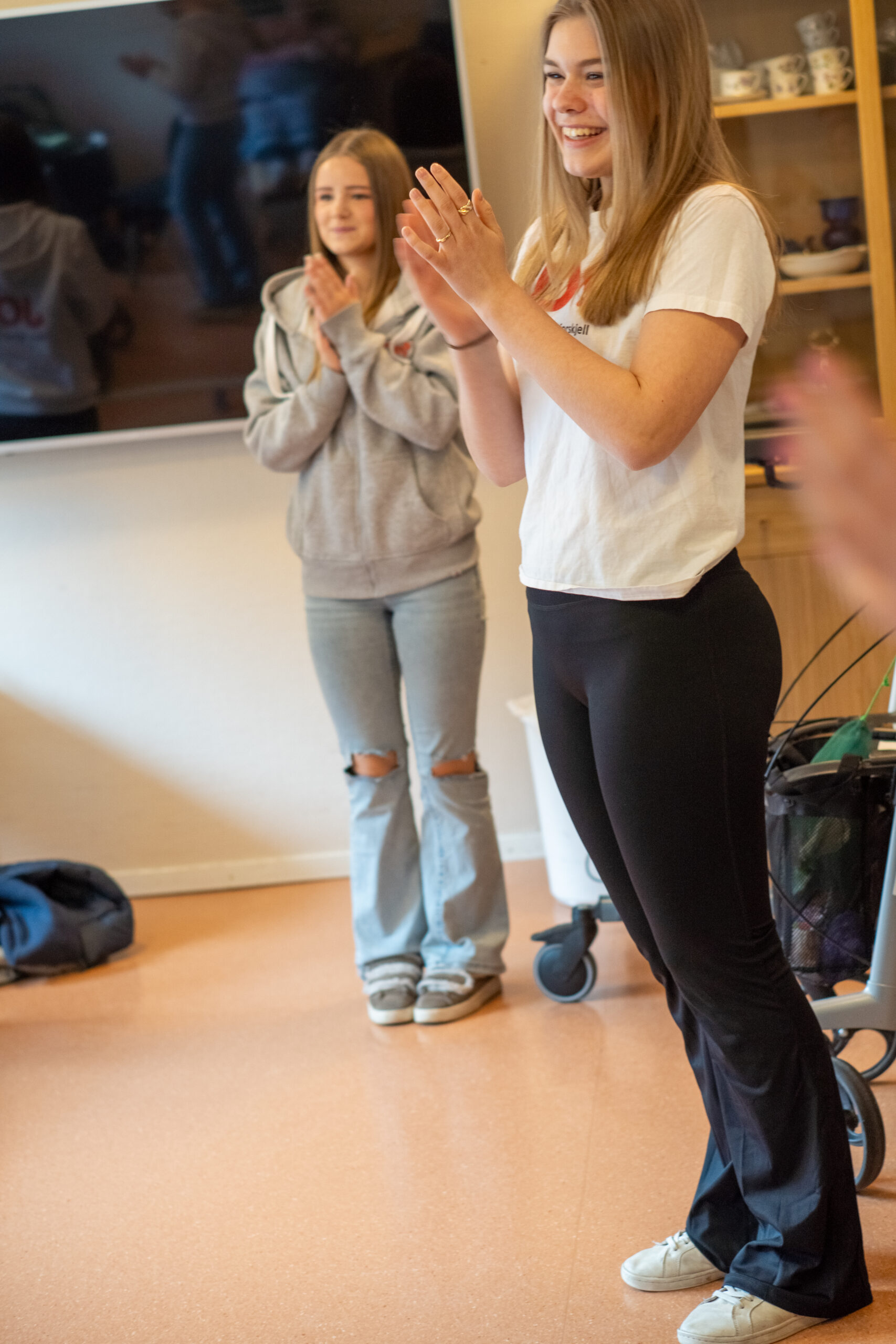 To unge kvinner står innendørs og smiler og klapper. Den ene har hvit t-skjorte og svarte bukser, den andre grå hettegenser og slitte jeans. I bakgrunnen skimtes hyller og en TV, som markerer en hyggelig stund sammen.