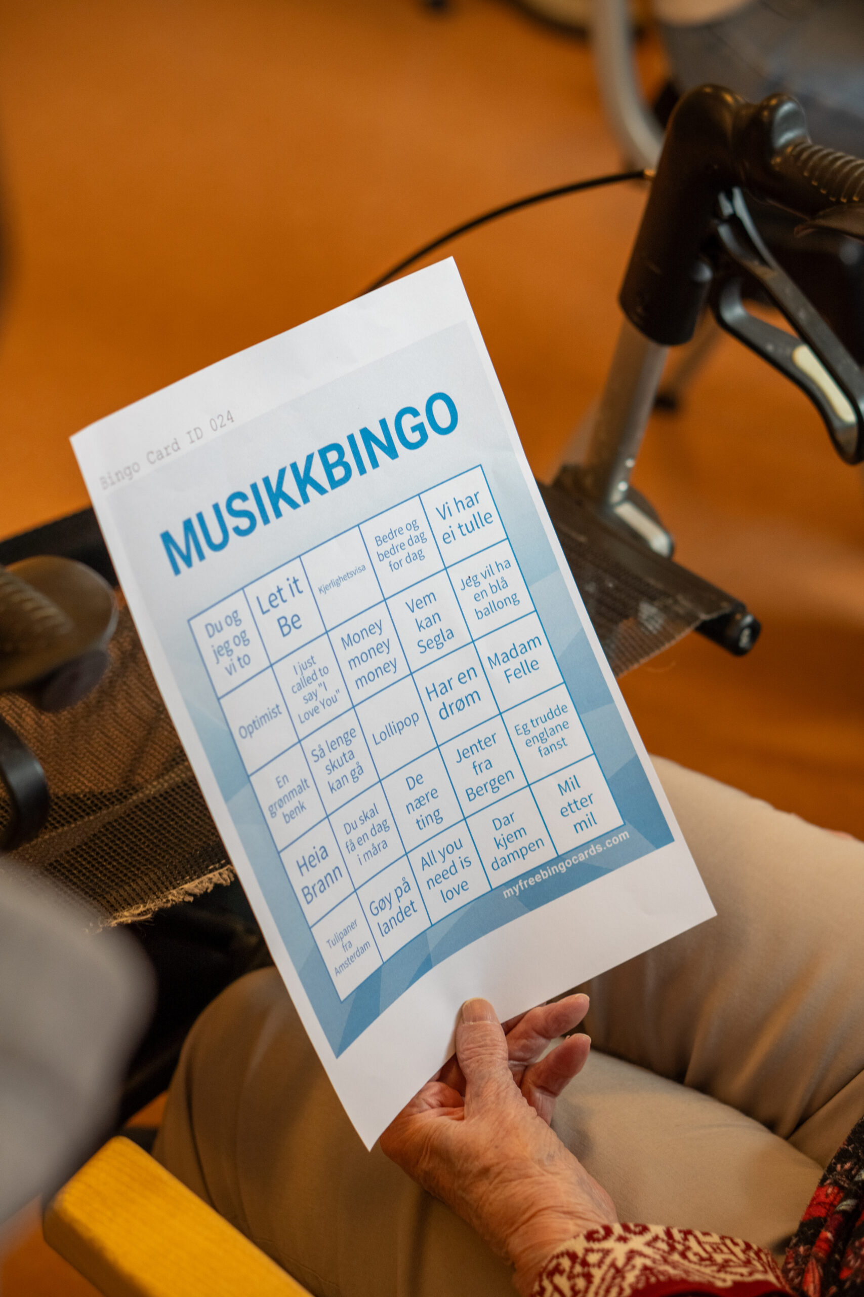 En person, eldre eller ung, holder et trykt ark med tittelen MUSIKKBINGO med sangtitler i et bingorutenett, sittende sammen ved siden av en rullator.