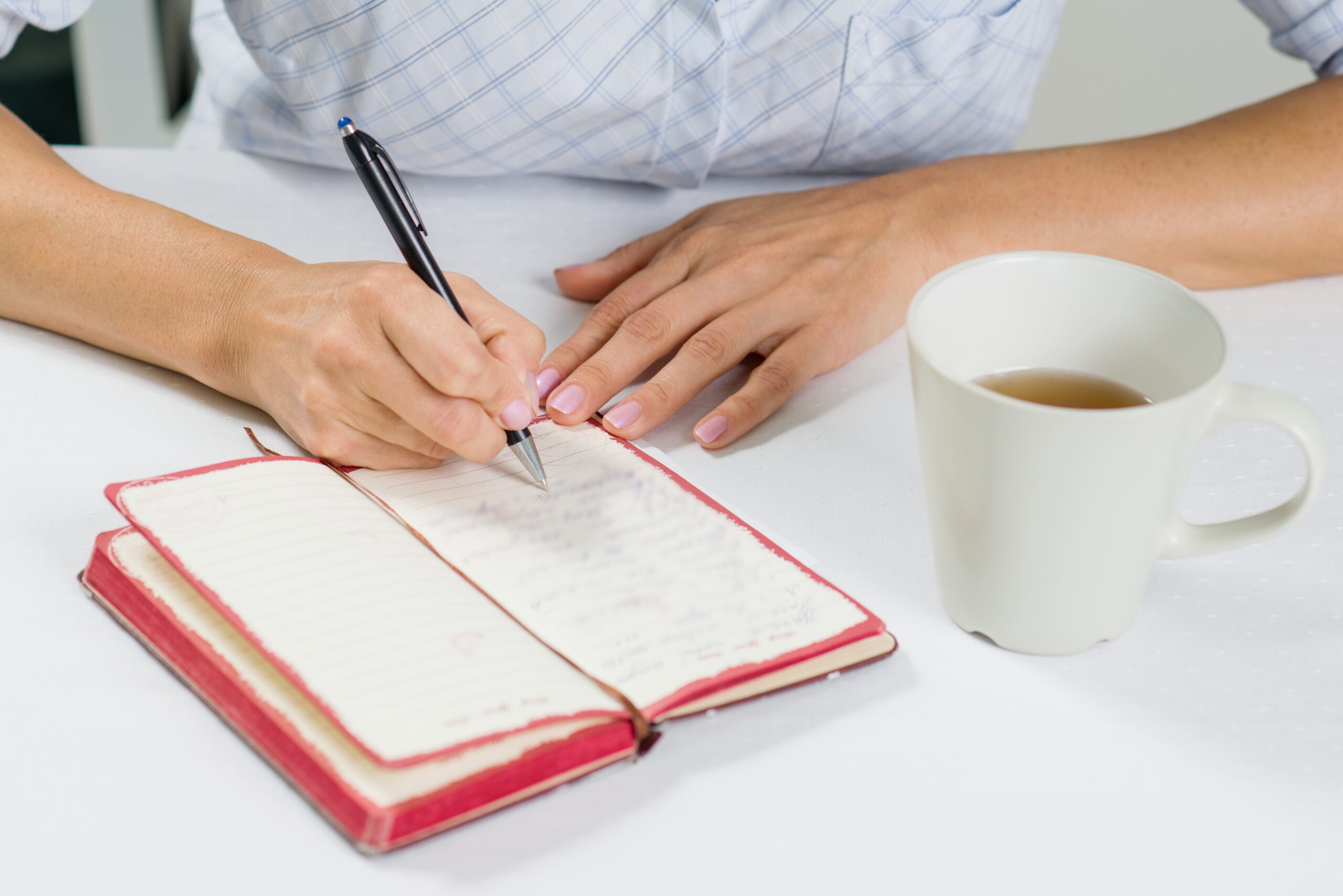 The hand of a woman with a pen writes in her diary. Next on the table is a cup of tea.