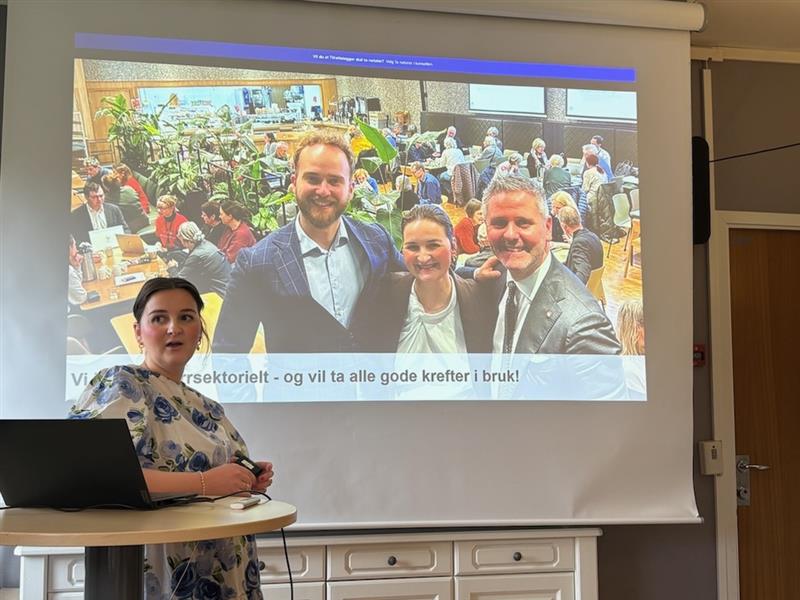 En kvinne står på et podium og presenterer et lysbilde som viser tre personer som poserer og smiler foran en gruppe under et møte.