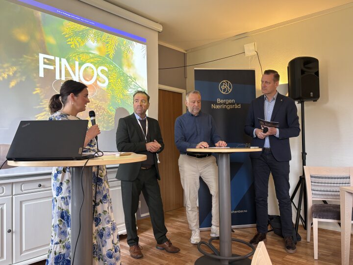Fire personer står og snakker på et frokostmøte innendørs, med en FINOS-presentasjonsskjerm og et banner fra Bergen Næringsråd synlig i bakgrunnen.