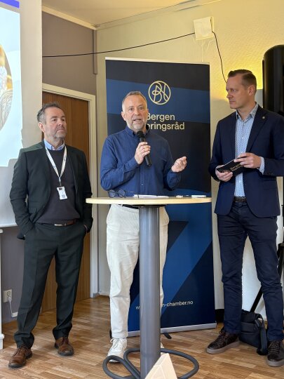 Tre menn i forretningsantrekk står ved et rundt bord under et frokostmøte, en av dem snakker i mikrofon, med et banner fra Bergen Næringsråd i bakgrunnen.