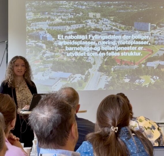 En kvinne holder foredrag på et frokostmøte i et konferanserom, med en projeksjon bak seg som viser tekst på norsk og et luftfoto av et boligområde.