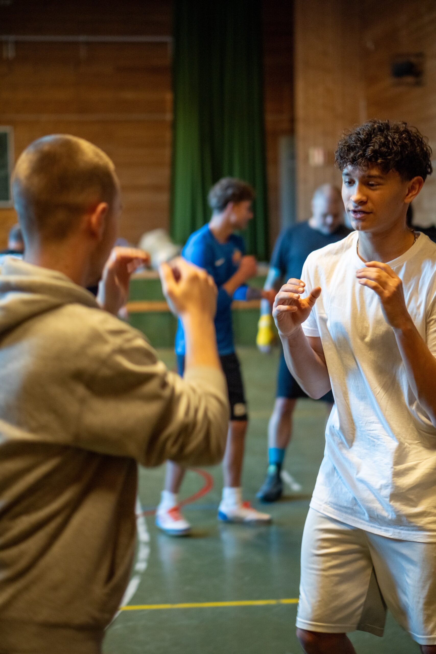 To unge menn står overfor hverandre i en gymsal og øver på en sparringsstilling, mens andre trener i bakgrunnen