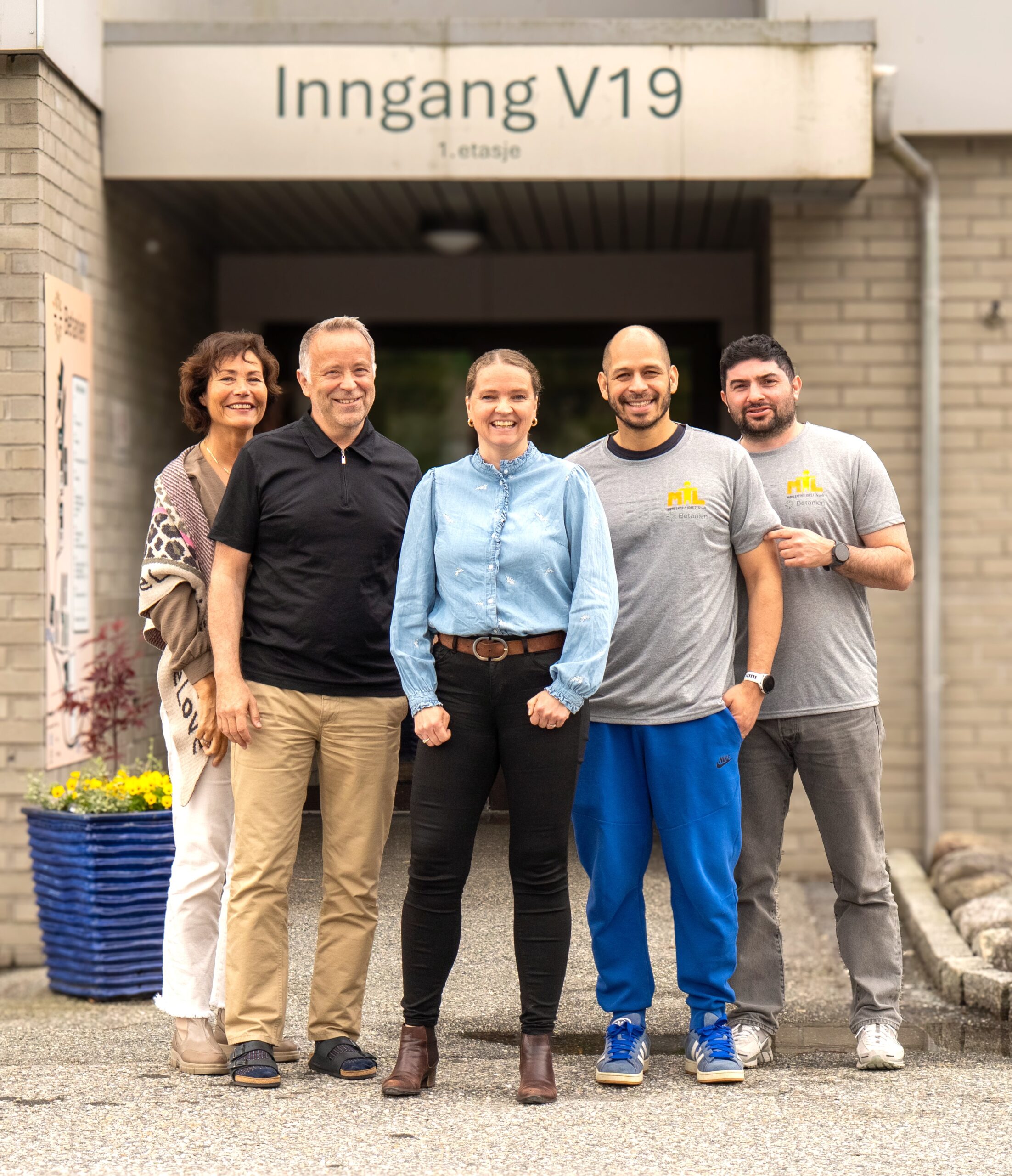 Fem personer står smilende foran inngangen til en bygning merket Inngang V19. Den lettkledde gruppen står sammen i fellesskap på en asfaltert gangvei.