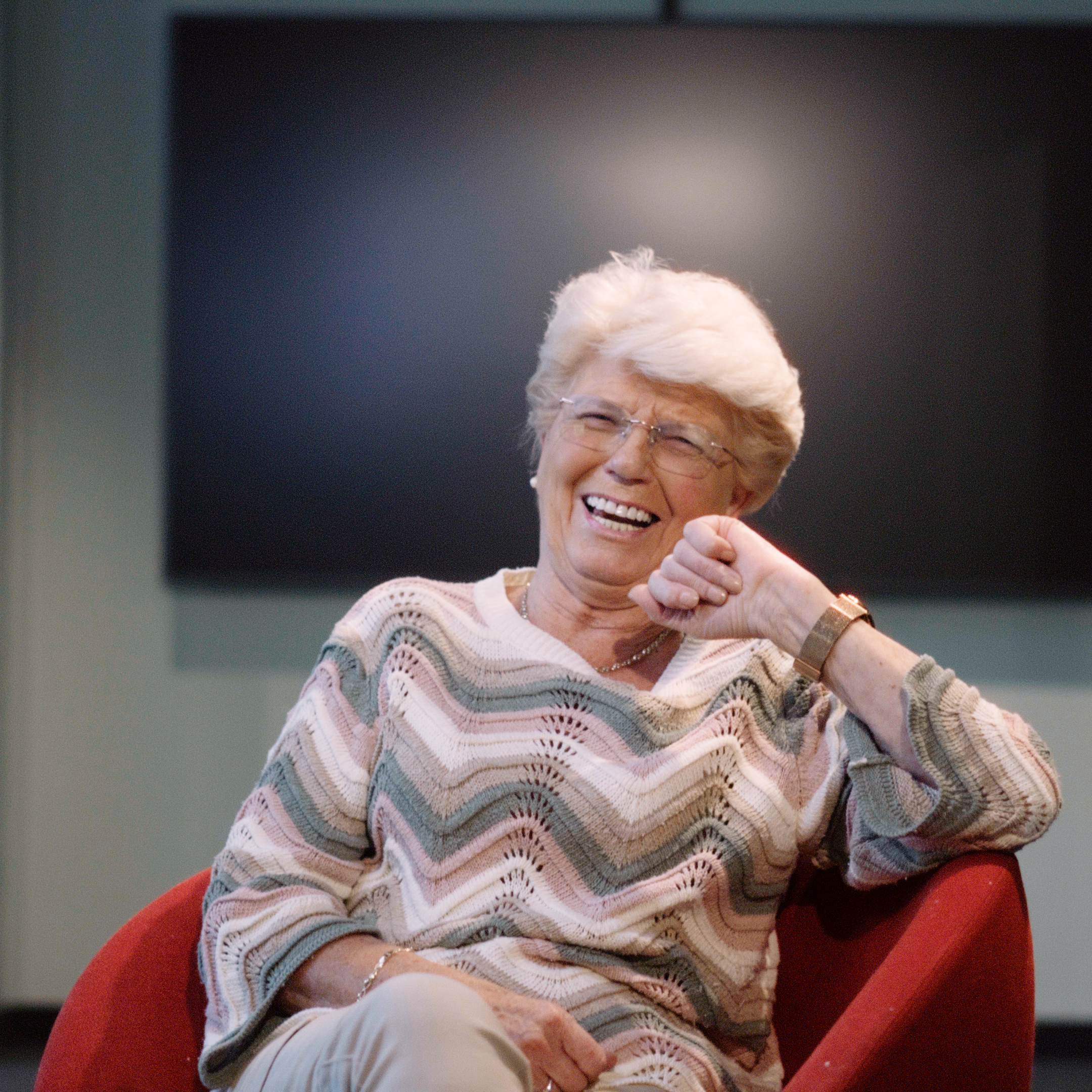 En 83 år gammel kvinne med kort, hvitt hår, iført briller og en mønstret genser, sitter i en rød stol og smiler fortsatt varmt. I bakgrunnen ser man en mørk, uklar skjerm